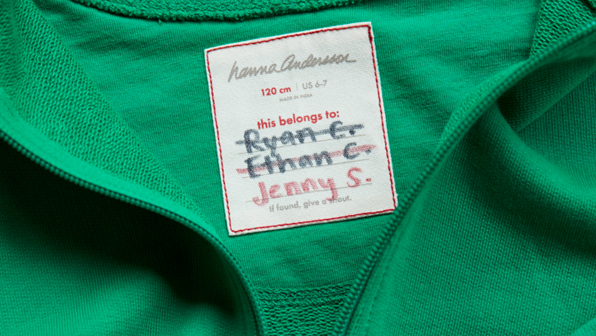 The tag of a Hanna Andersson hoodie with two names crossed out and one not crossed out indicating that this article of clothing has been handed down over generations